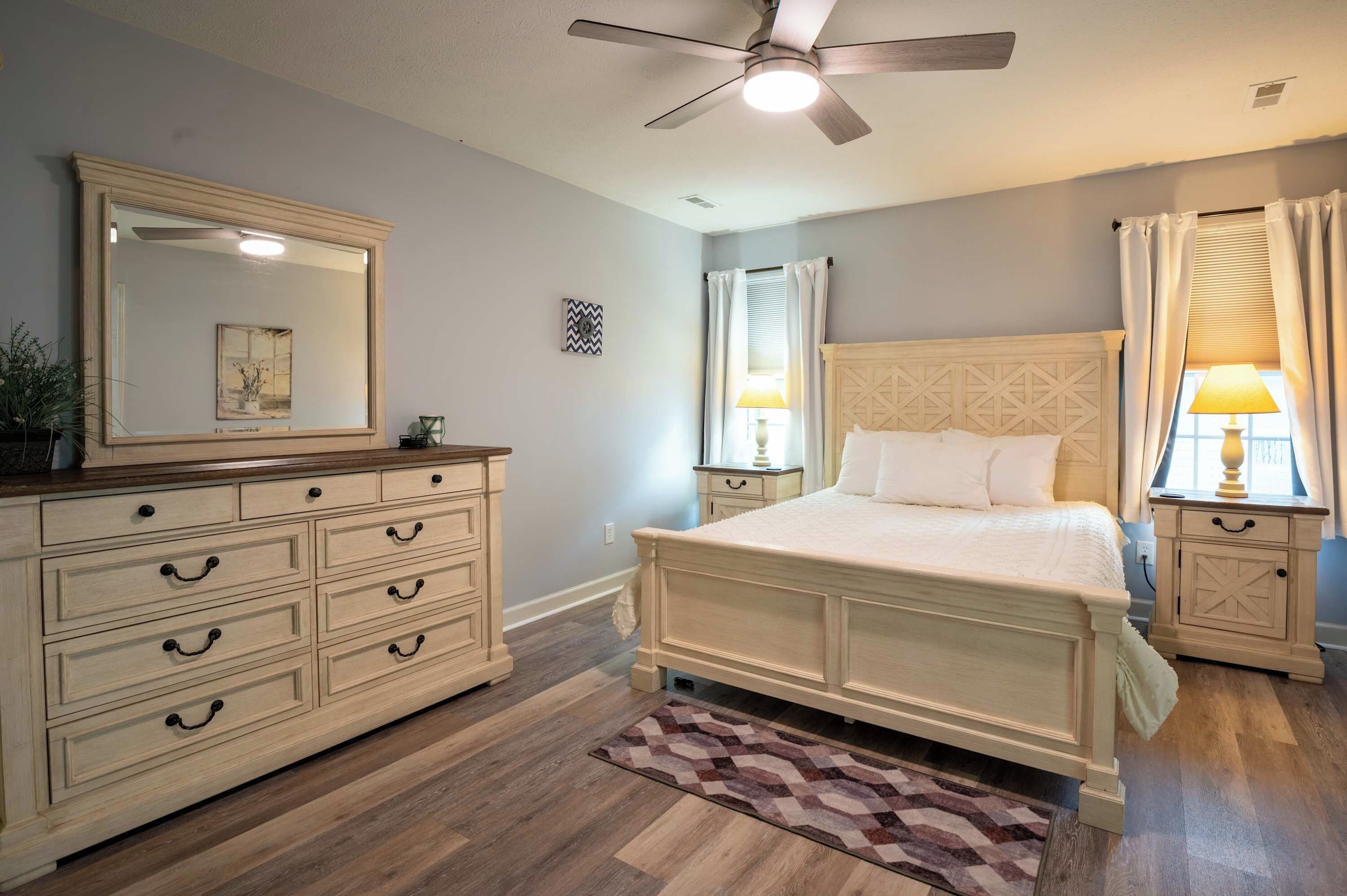 A bedroom with a bed, a dresser, a nightstand, a ceiling fan, and a window with curtains.