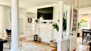 A spacious living room with a fireplace, a television mounted above it, and a dining area with a table and chairs. - Maxson House Mystic – Easy Walk to Downtown (Mystic)