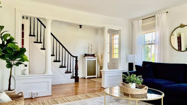 A living room with a staircase, a dark blue sofa, a round coffee table, and a large mirror. - Maxson House Mystic – Easy Walk to Downtown (Mystic)