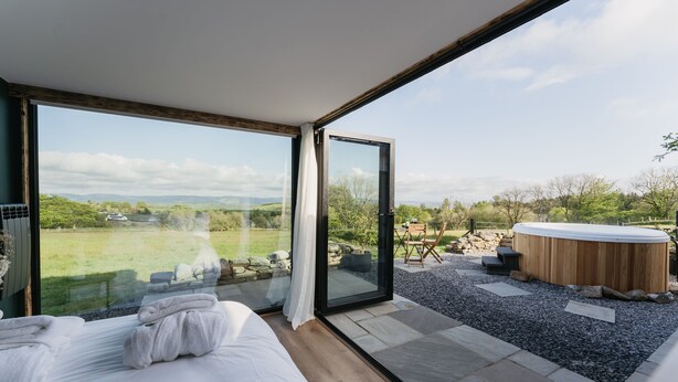 A modern bedroom with a large glass door leading to a terrace with a hot tub and a scenic view. - Dyffryn Ceri - luxury converted shipping container (Penuwch)