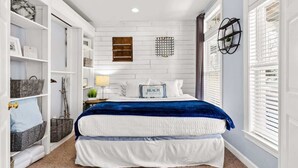 Un dormitorio con una cama blanca, una manta azul y un cabecero blanco. Hay un estante de madera pequeño y una lámpara de pared.