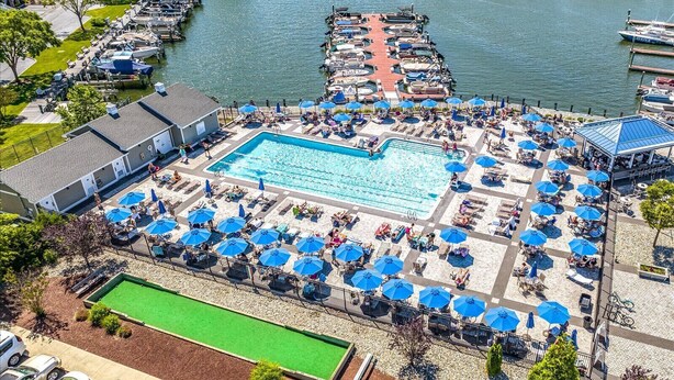 An aerial view of a resort with a large swimming pool surrounded by sun loungers and umbrellas, a dock with boats, and a river in the background. - Ocean Pines Hideaway (Ocean Pines)