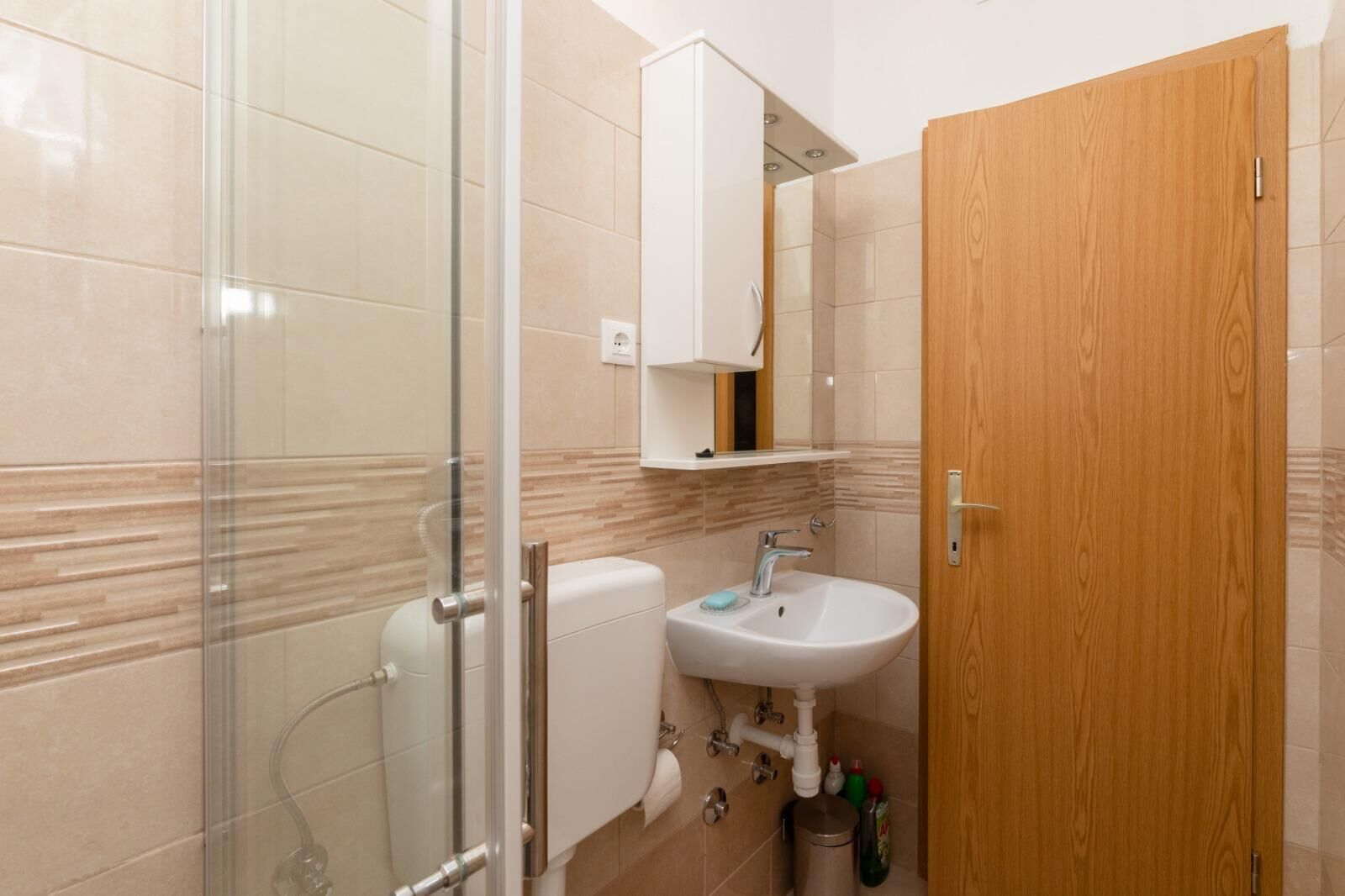 A bathroom with a glass shower enclosure, a white sink, and a wooden door.