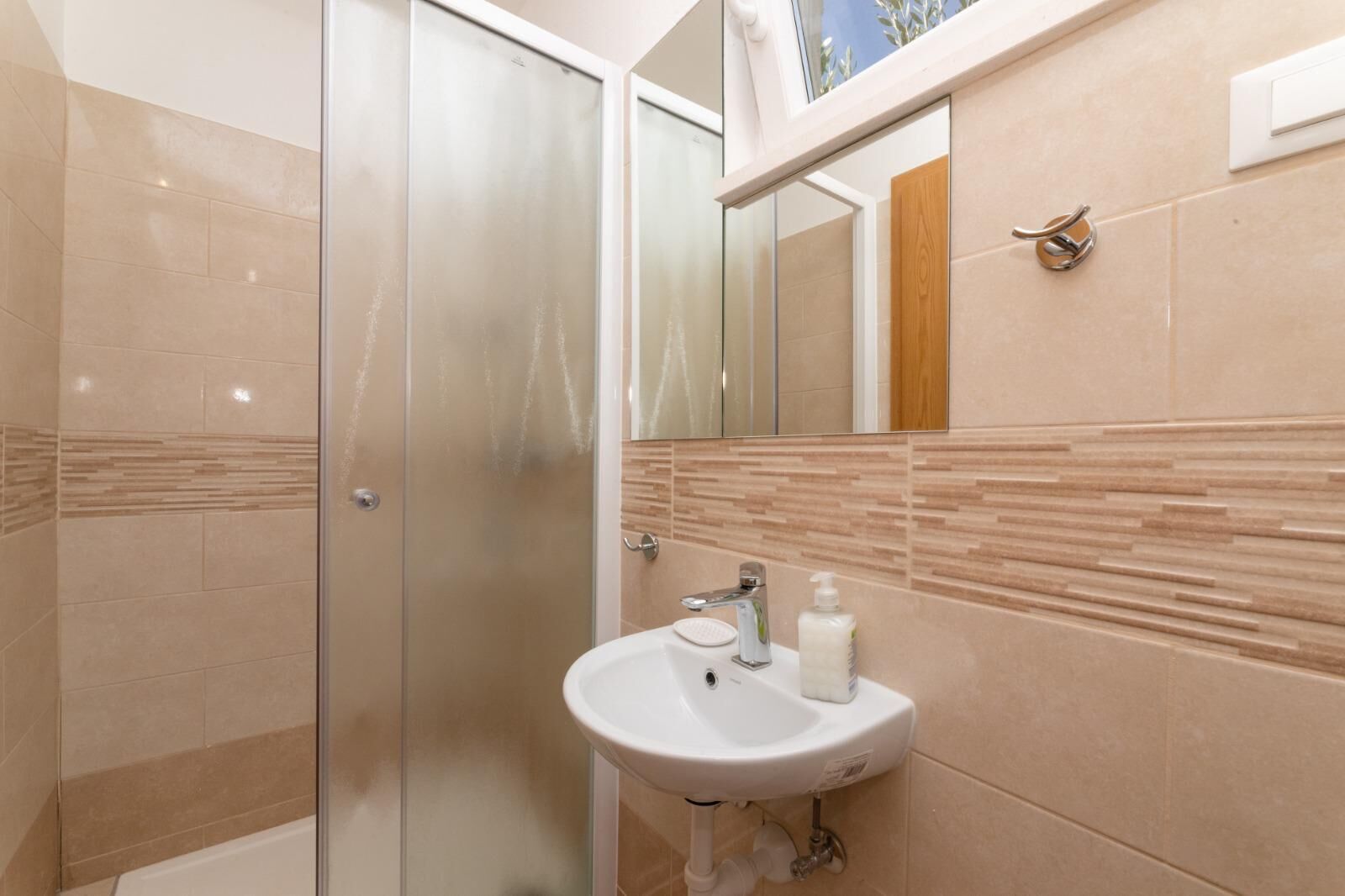 Een badkamer met een douchecabine van glas, een wastafel met spiegel en beige betegelde wanden.