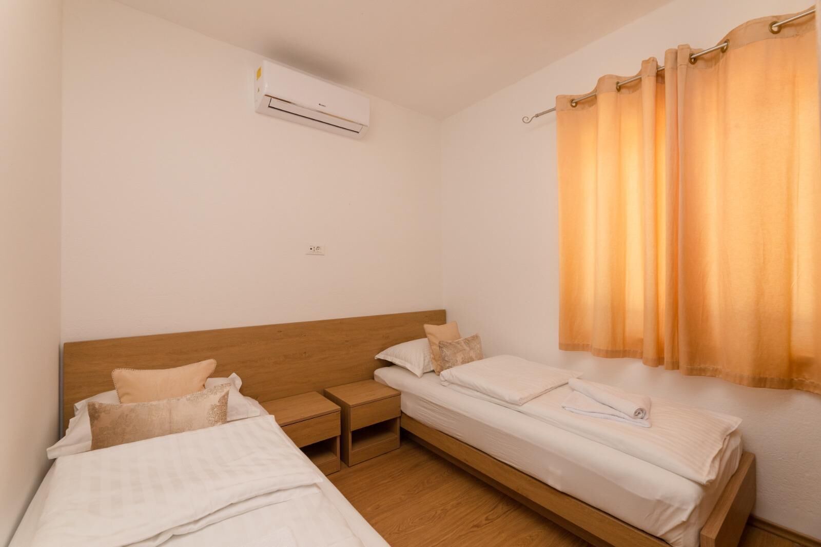 Quarto com duas camas de solteiro, cabeceira de madeira, mesa de cabeceira, ar condicionado e janela com cortinas.