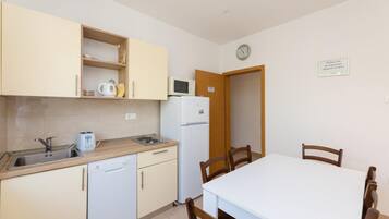 Cozinha compacta com pia, torradeira, chaleira e micro-ondas. Há uma mesa de jantar com cadeiras e uma porta que dá para outro quarto.
