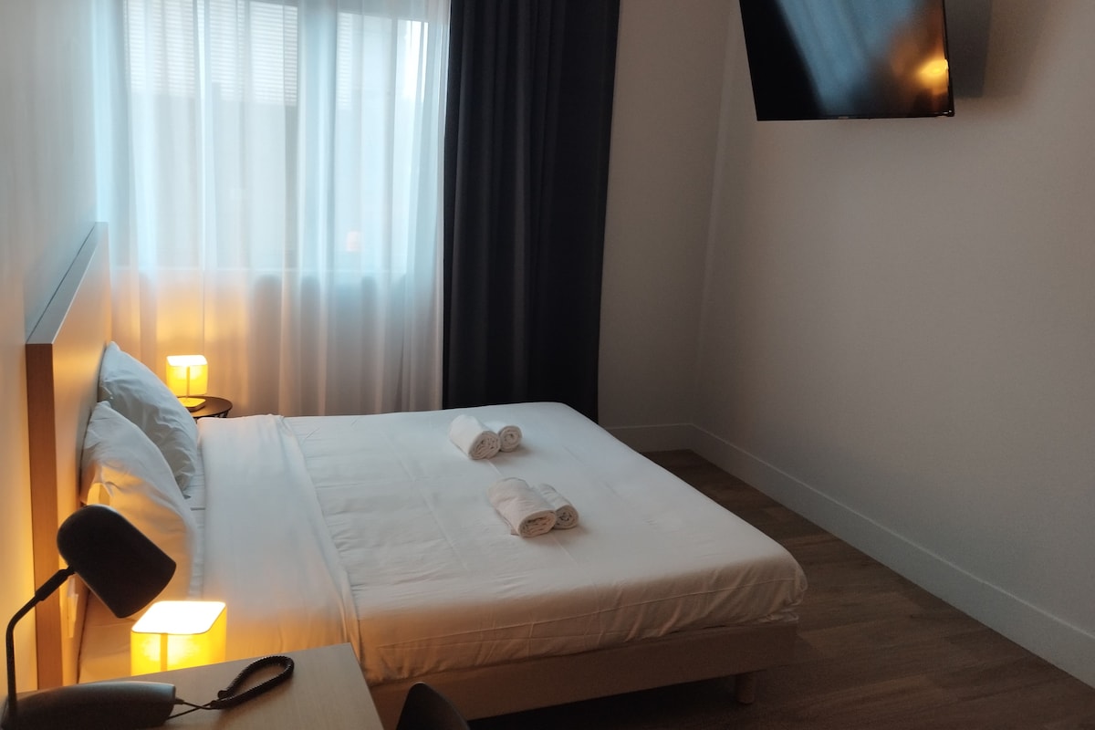 Ett hotellrum med en säng, ett skrivbord, en lampa, en tv som är monterad på väggen och ett fönster med gardiner.