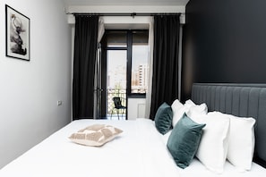 Quarto de hotel moderno com uma cama grande, cortinas escuras, vista para a cidade e uma obra de arte emoldurada na parede.