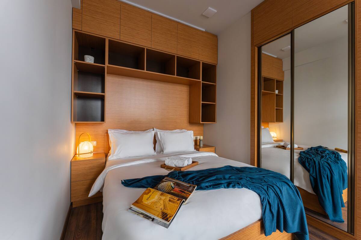 A modern hotel room with a large bed, wooden headboard, built-in shelves, a bedside table with a lamp, and a large mirror.
