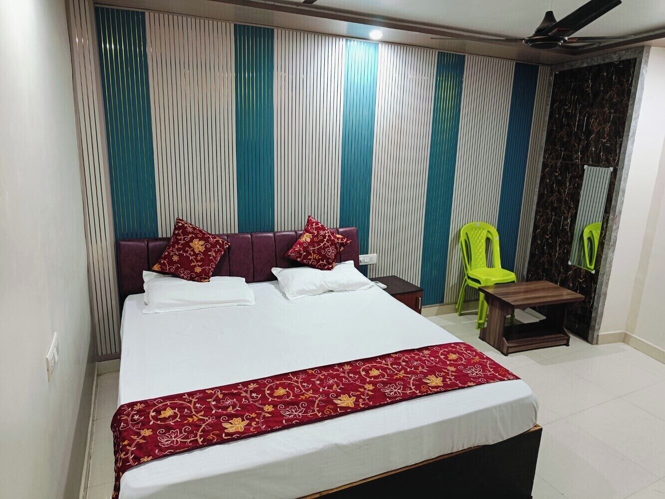 Quarto de hotel com uma cama grande, uma mesinha de cabeceira de madeira, uma cadeira verde e uma pequena mesa.