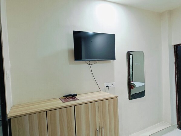 Ein Zimmer mit an der Wand montiertem Fernseher, einem Spiegel und einer Holzkassette mit Fernbedienung darauf.