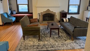 Un salon avec une cheminée, un fauteuil bleu, un canapé gris et un tapis à motifs.