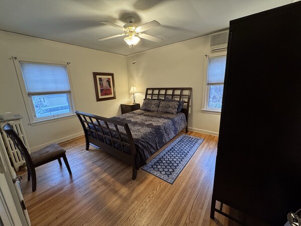 A bedroom with a bed, a nightstand, a chair, a ceiling fan, and two windows.