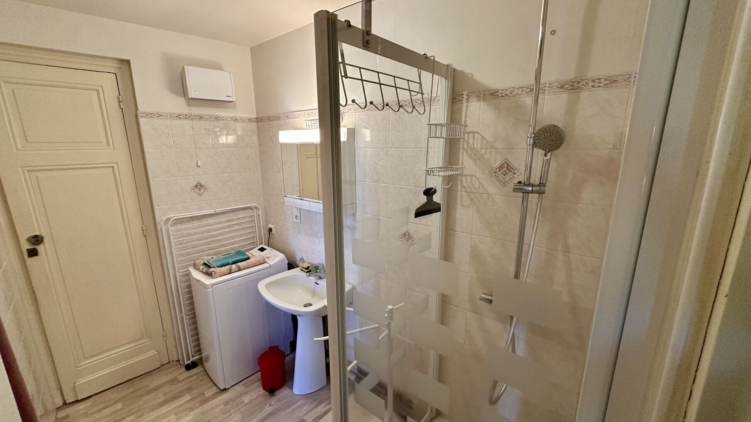 A bathroom with a glass shower enclosure, a sink, a washing machine, and a door with a towel rack.
