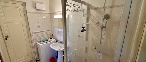 A bathroom with a glass shower enclosure, a sink, a washing machine, and a door with a towel rack.