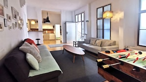 Un salon moderne avec un canapé, une table basse et un baby-foot. On aperçoit une cuisine à l’arrière-plan, avec des placards et un luminaire suspendu.
