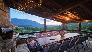 An outdoor dining area with a stone floor, wooden table, and chairs, overlooking a mountainous landscape. - Authentic Dalmatian Hillside Escape With Pool, Sauna & BBQ (Sestanovac)
