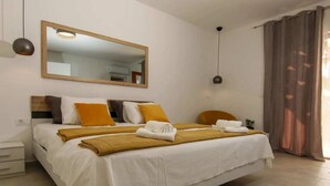 Un lit bien fait, avec une literie blanche et moutarde, une tête de lit en bois et un grand miroir au-dessus. Il y a deux tables de chevet, l’une avec une lampe et l’autre avec une prise de courant.