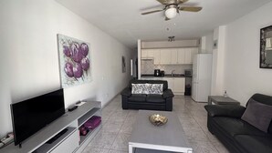 A modern living room with a flat-screen TV, a ceiling fan, and a kitchen in the background.
