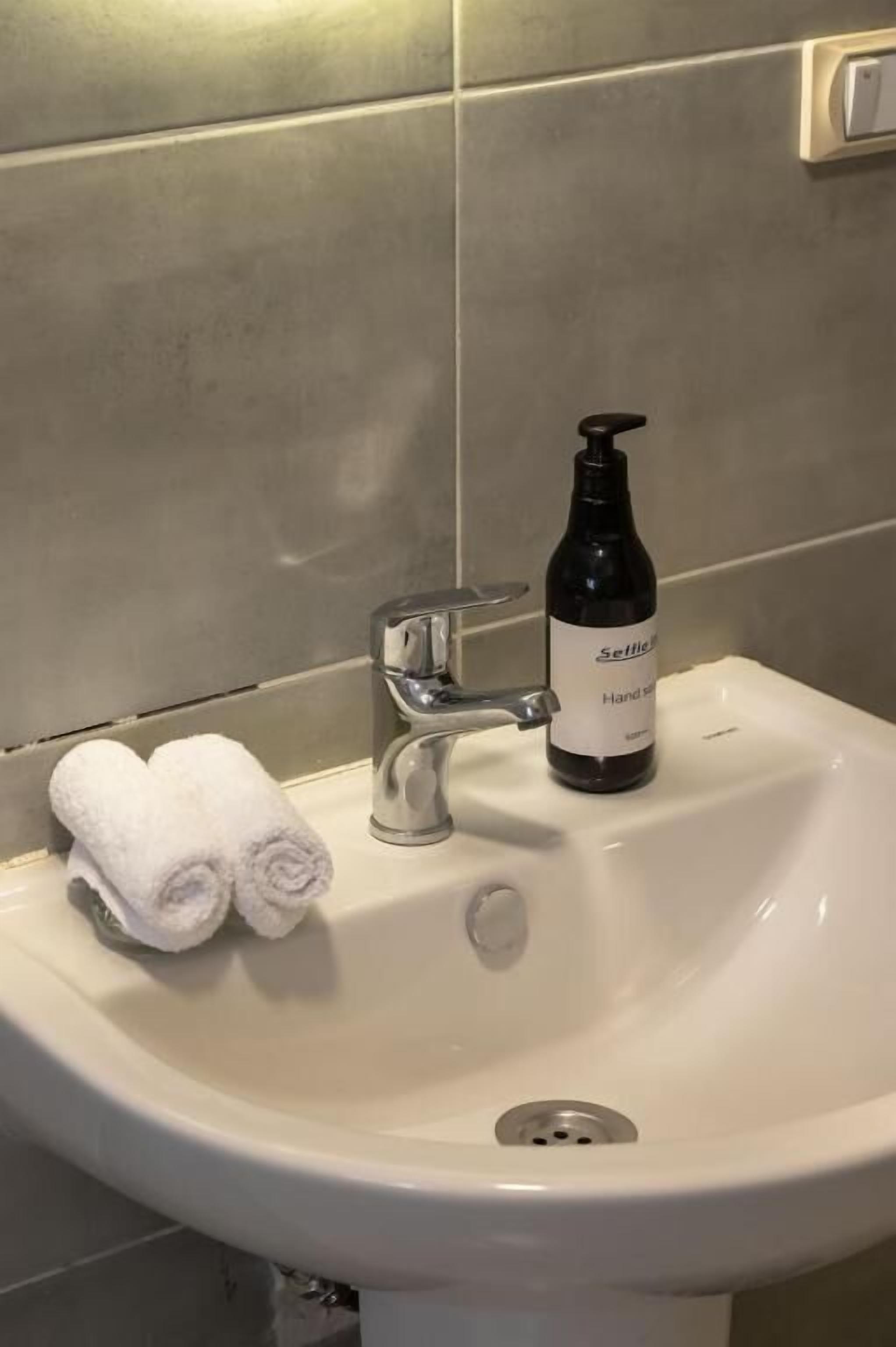 A white bathroom sink with a silver faucet, a bottle of hand soap, and two rolled towels on the sink.