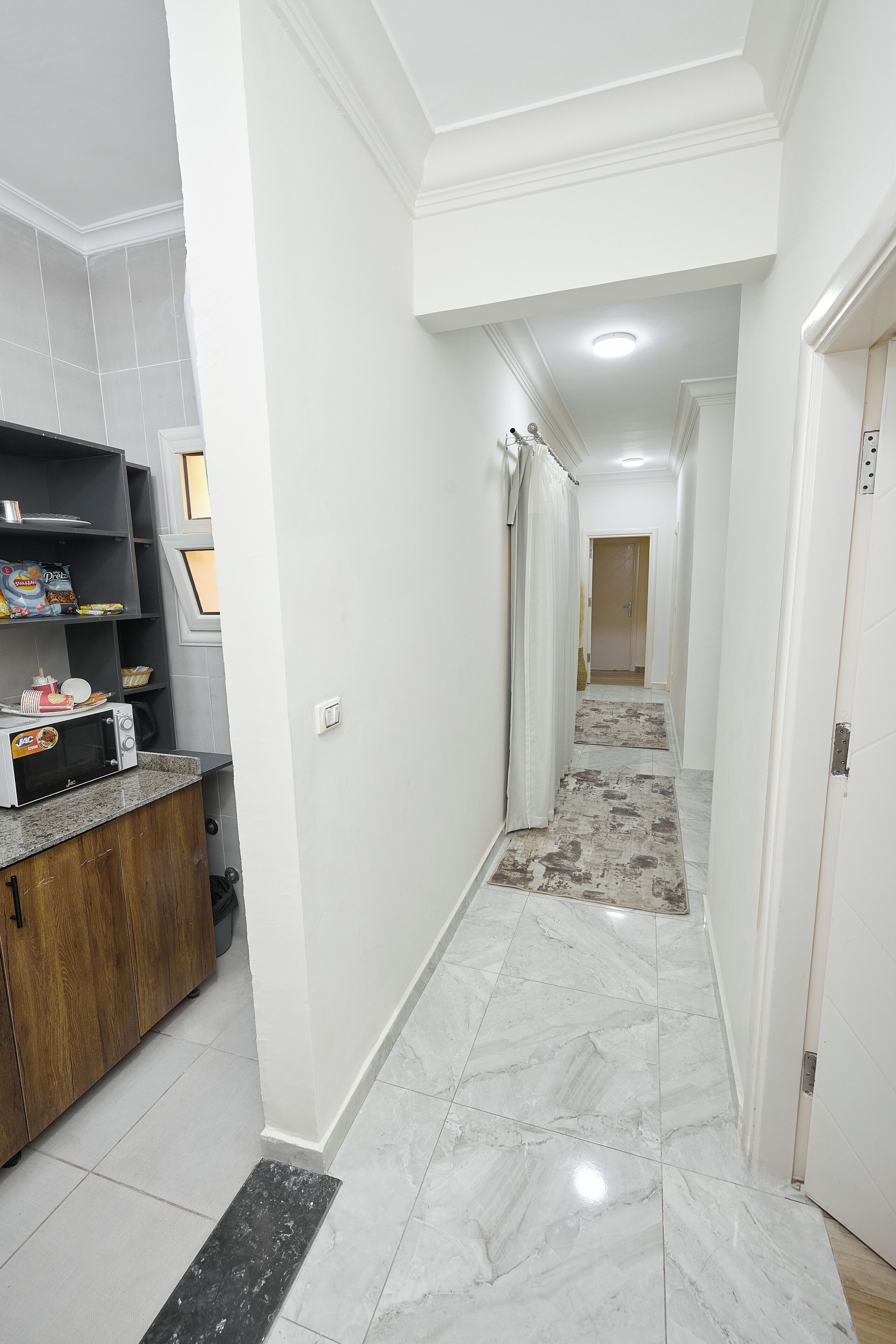 Un pasillo con una zona de cocina a la izquierda, que incluye un microondas y armarios. El piso está cubierto de baldosas y las paredes son blancas.