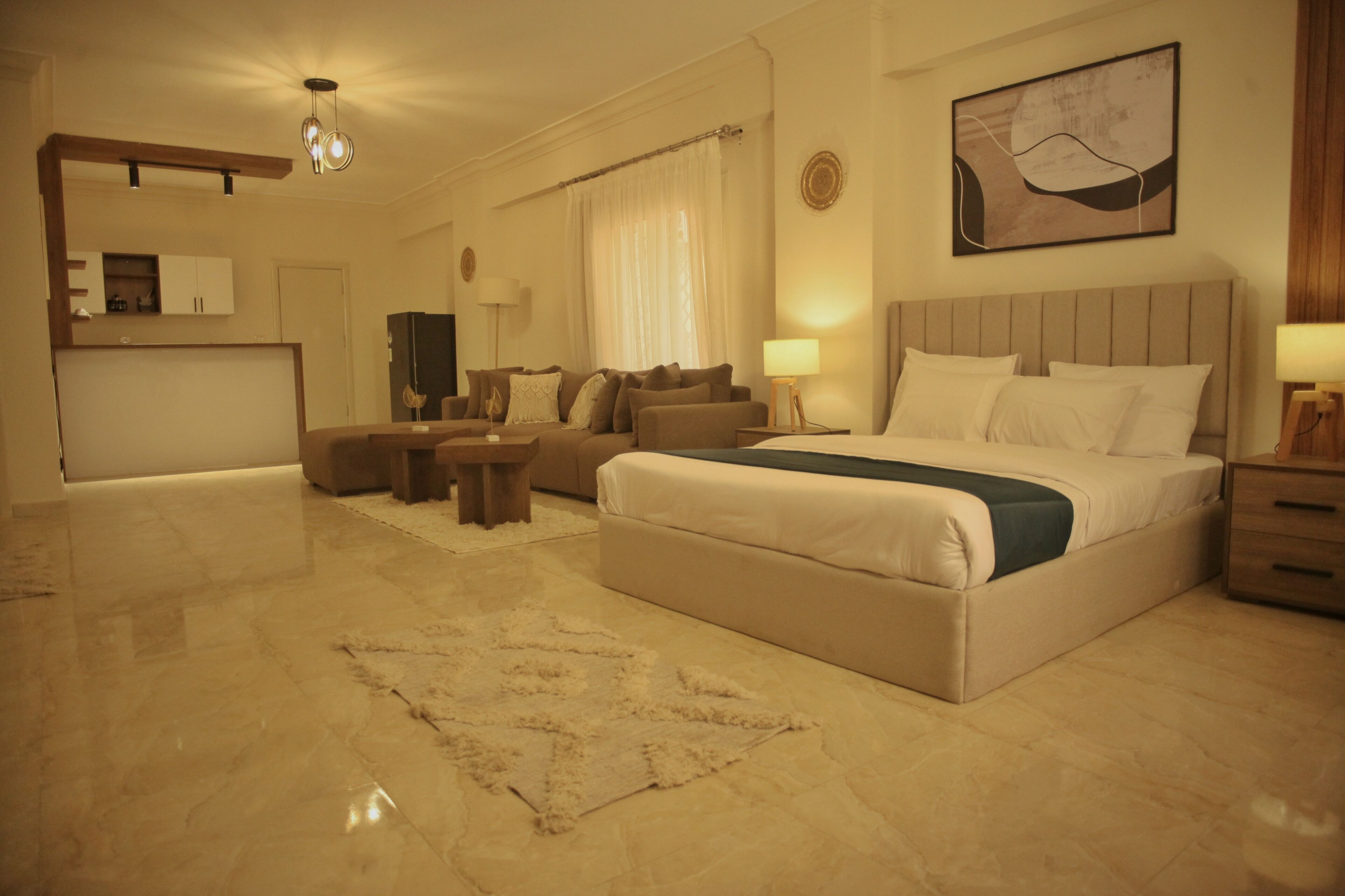 A modern hotel room with a large bed, a sofa, a small table, a TV, and a kitchen area.