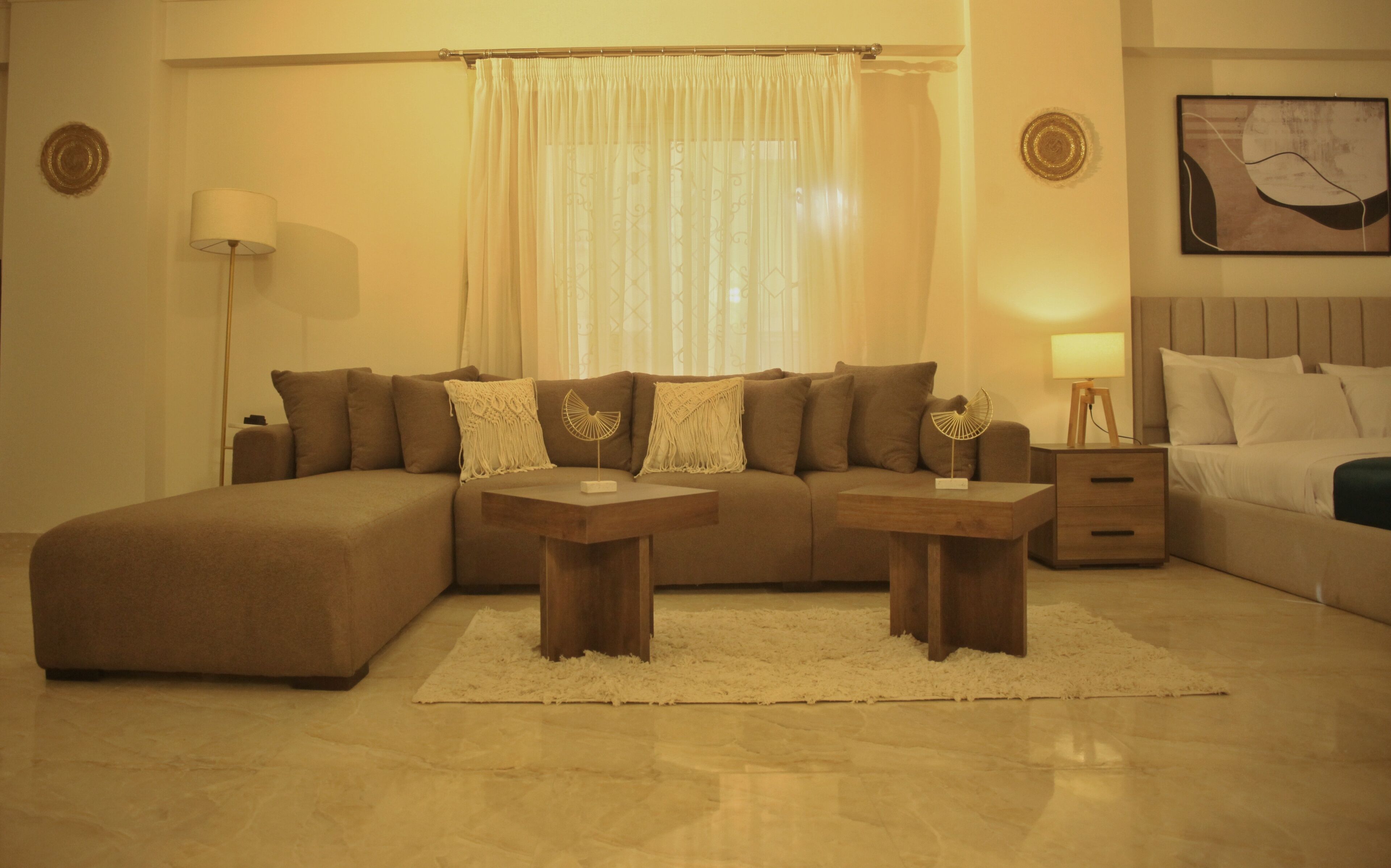 A modern living room with a sectional sofa, two wooden side tables, a white rug, and a bed with a headboard.