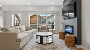 Un salon moderne comprenant un canapé, un fauteuil, une table basse et des poufs, avec une grande télévision et de grandes fenêtres donnant sur l’extérieur.