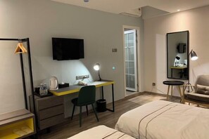 A hotel room with a bed, a desk with a chair, a flat-screen TV, a mirror, and a small table with a lamp.