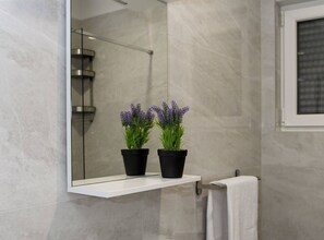 Ein Badezimmer mit Marmorwand, einer Duschkabine aus Glas und zwei Lavendelpflanzen in Töpfen auf einem weißen Regal.