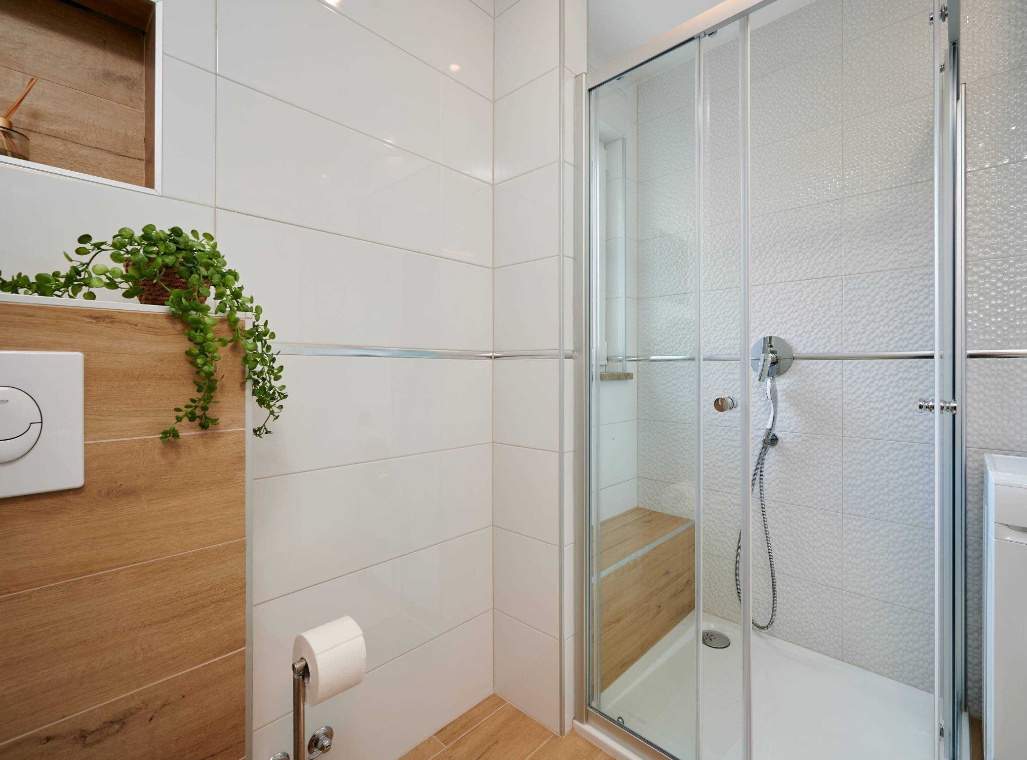 A modern bathroom with white tiles, a glass shower enclosure, a wooden vanity, and a green plant.