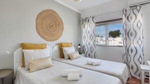 Habitación de hotel con dos camas, un tapete redondo tejido y vistas a edificios a través de una ventana con cortinas estampadas.