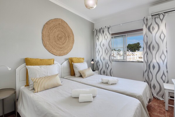 Une chambre d’hôtel avec deux lits, un tapis rond en fibres tressées et une vue sur les bâtiments par une fenêtre ornée de rideaux à motifs.