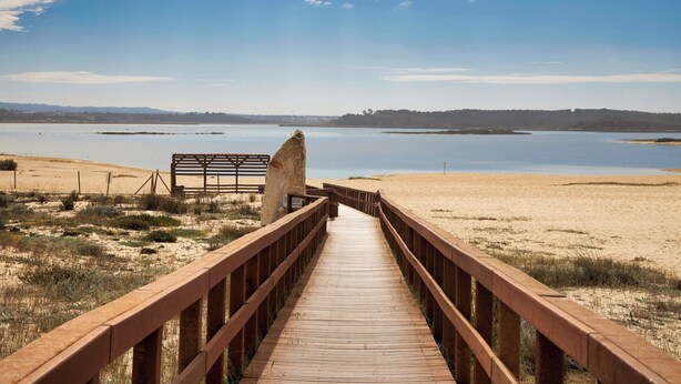 Un paseo marítimo de madera que conduce a una zona de playa arenosa, con vista despejada a una masa de agua tranquila y colinas lejanas.