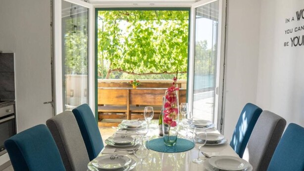 A dining area with a table set for four, a glass vase with flowers, and a view of greenery outside.