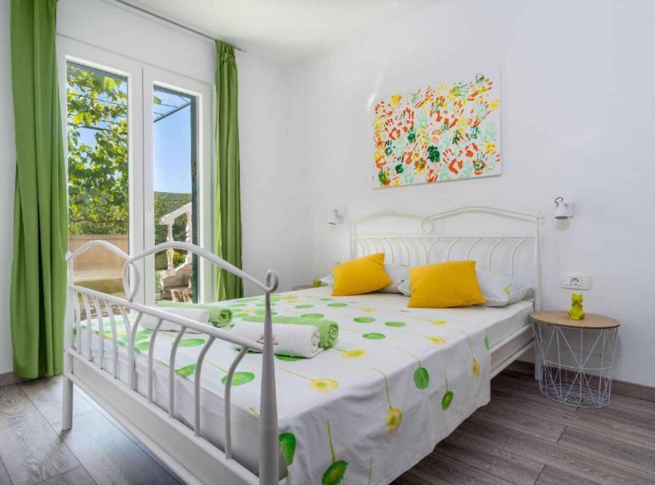 A bedroom with a bed, green curtains, a window, a painting, and a small round table with a yellow object.