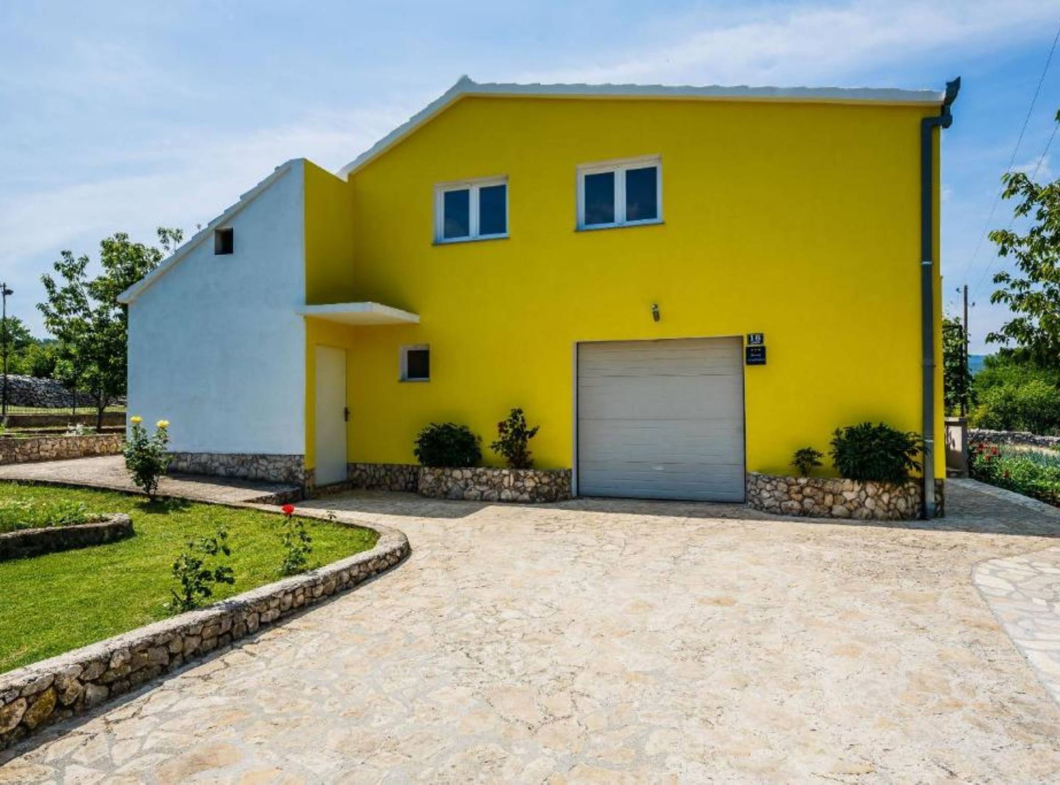 A two-story yellow building with a white section on the left, a garage door, and a small garden area with a stone border.
