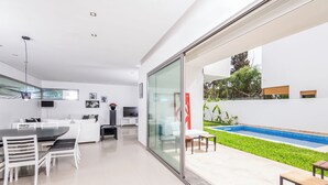 Eine moderne Küche mit weißen Schränken, ein Essbereich mit Tisch und Stühlen und eine Glastür, die zu einem Poolbereich mit einer kleinen Terrasse führt.