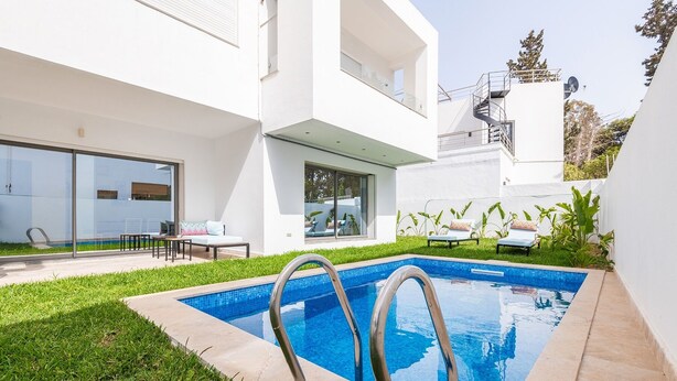 Ein modernes Haus mit Swimmingpool, einer Terrasse mit Liegestühlen und einer Glastür zu einem überdachten Bereich.