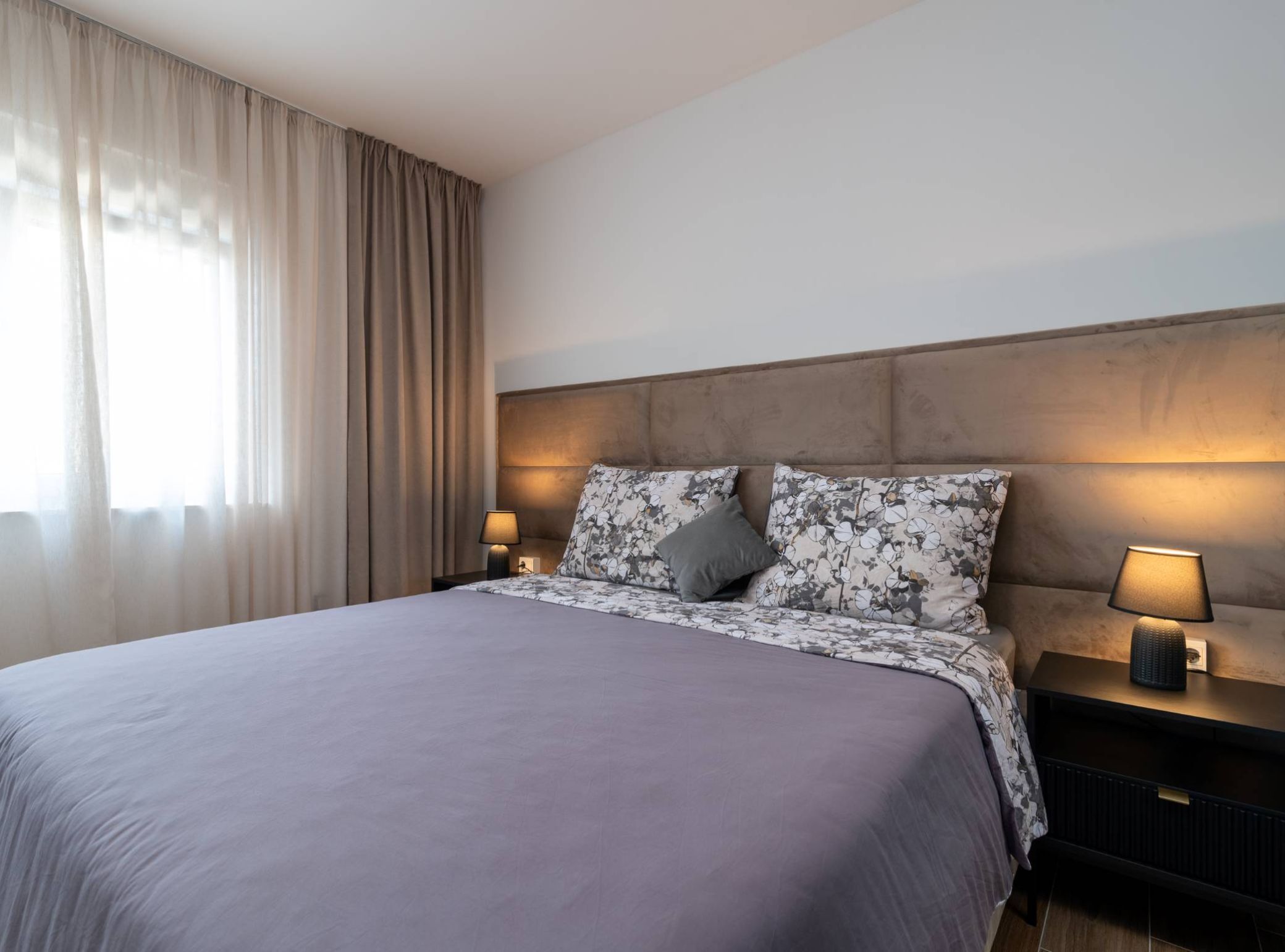 A hotel room with a large bed, two bedside tables with lamps, a headboard, and a window with curtains.