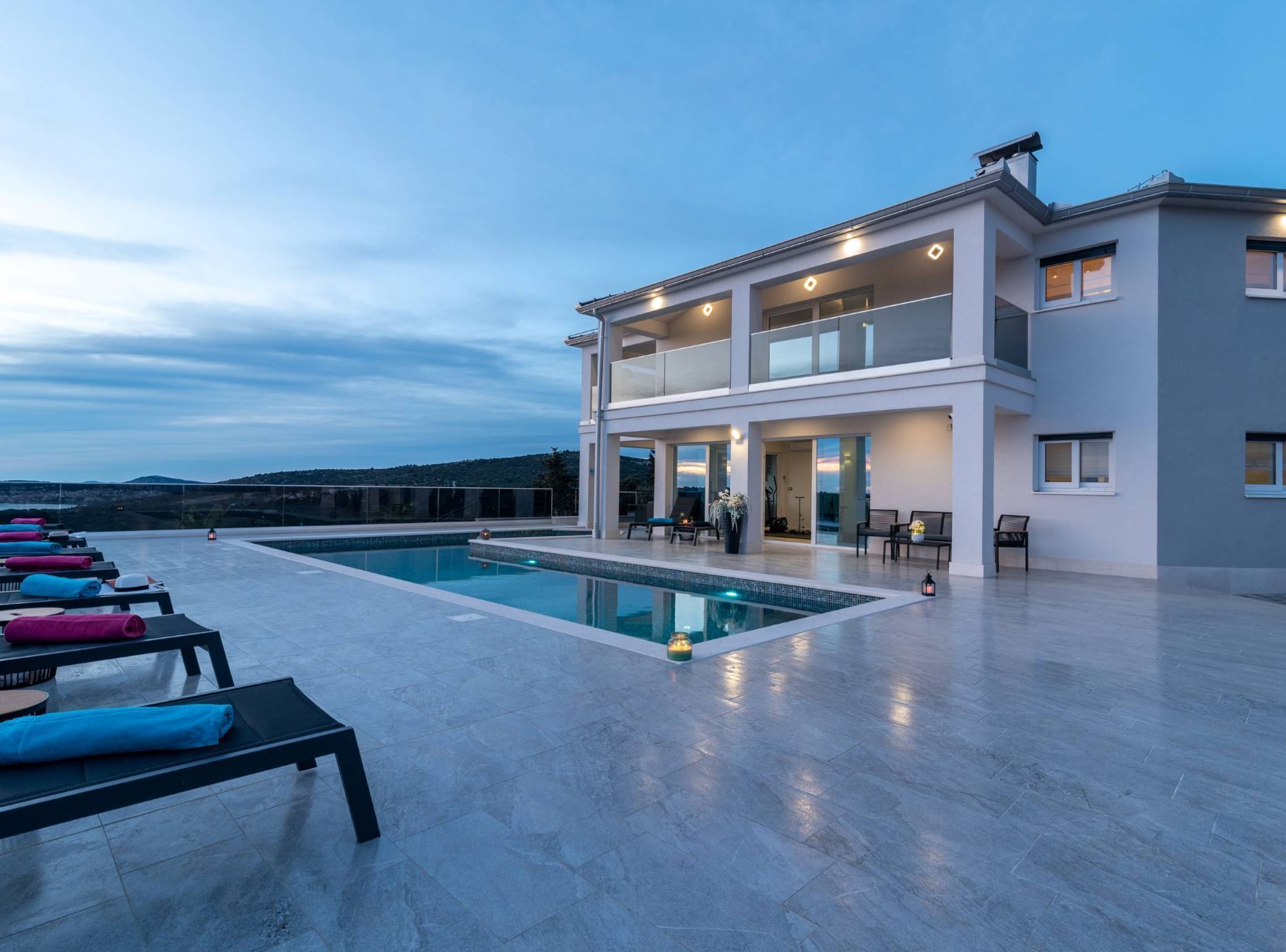 A modern two-story building with a rooftop pool area, lounge chairs, and a scenic view.