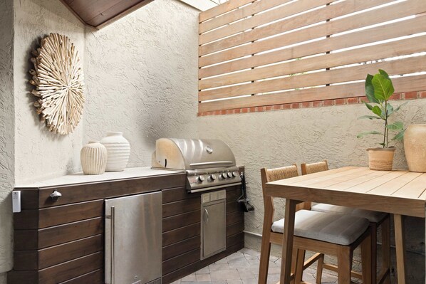A compact outdoor kitchen with a built-in grill, a refrigerator, and a wooden dining table with chairs.