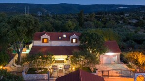 Une maison à deux étages, avec un toit recouvert de tuiles rouges, entourée d’arbres et de collines.