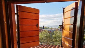 Une fenêtre avec des volets en bois est ouverte, dévoilant une vue sur un toit avec des tuiles et quelques plantes.