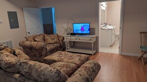 A living room with a floral sofa, a TV on a white console table, and a bathroom visible through an open door. -  1-bdrm house with fireplace, AC and WiFi with 20 acres of  trees scenic deck. (Kingston)