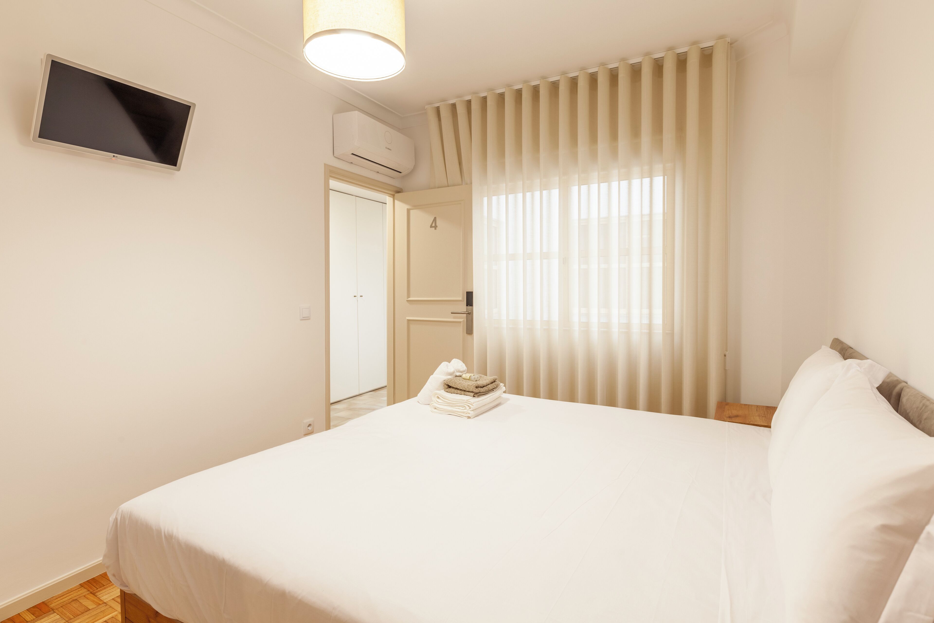 A hotel room with a bed, a TV mounted on the wall, an air conditioning unit, and a door leading to another room.