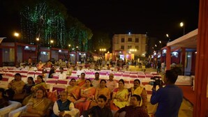 Evento ao ar livre com convidados sentados e palco, cercado por tendas e árvores.