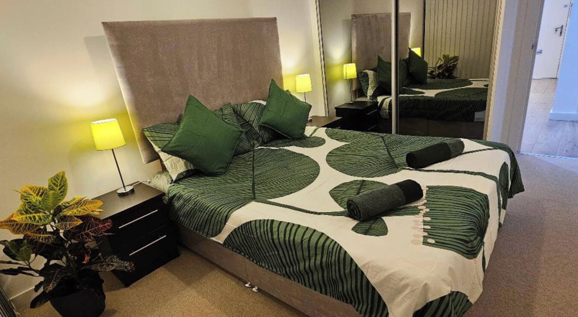 A neatly made bed with green and white bedding, a bedside lamp, and a potted plant.