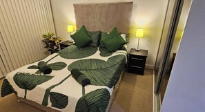 Un lit bien fait, recouvert d’une couvre-lit à motifs de feuilles, deux tables de chevet avec des lampes et une plante en pot sur l’une d’elles.