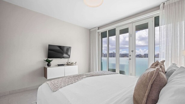 A modern bedroom with a large bed, a TV on a white cabinet, and a city view through large glass doors. - Wave Front Escape | Trendy Studio in Condado (San Juan)